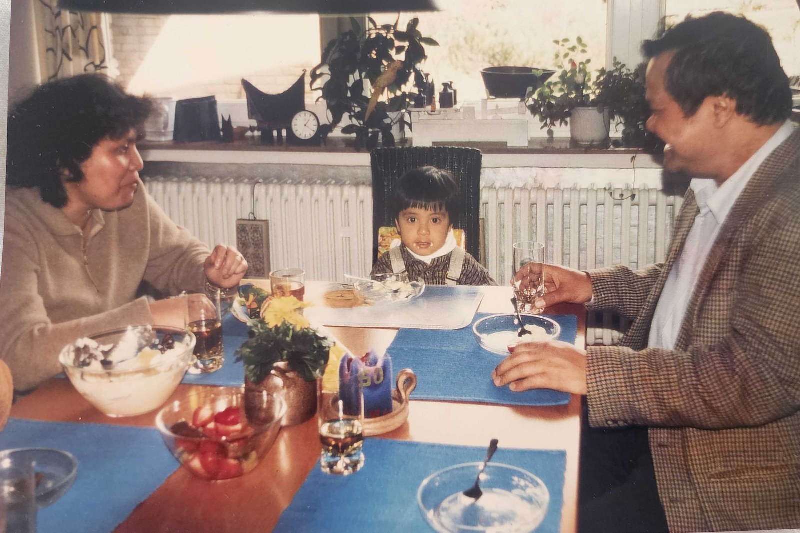 My mother and father at either end of a table with me in the middle in a high chair, bowls of dessert in front of us