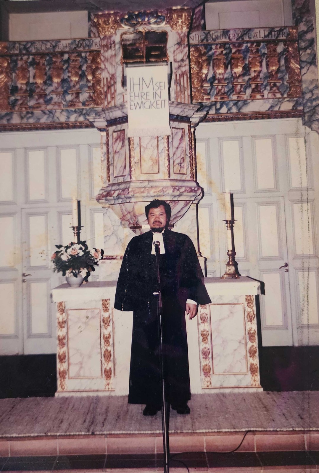 My dad in black clerical robes standing at the altar of a German church, a banner above him reading Ihm sei Ehre in Ewigkeit