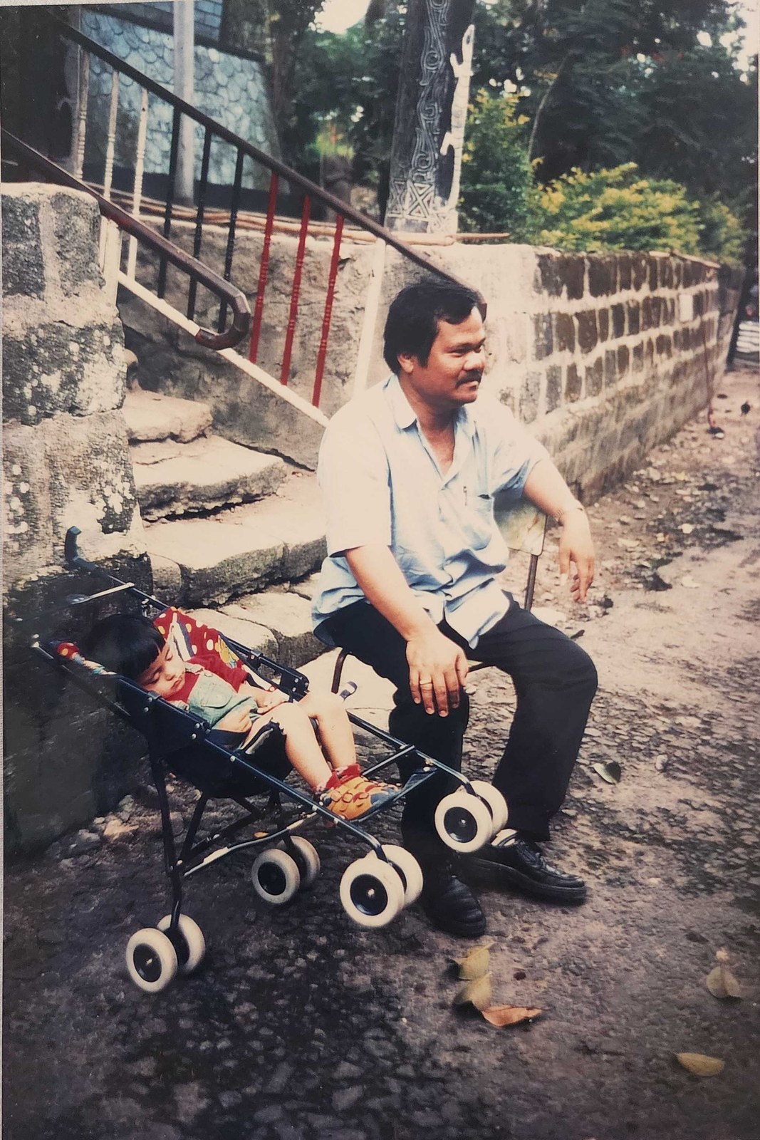 My dad sitting on a low wall beside me, asleep in a pushchair, with a carved wooden post behind us