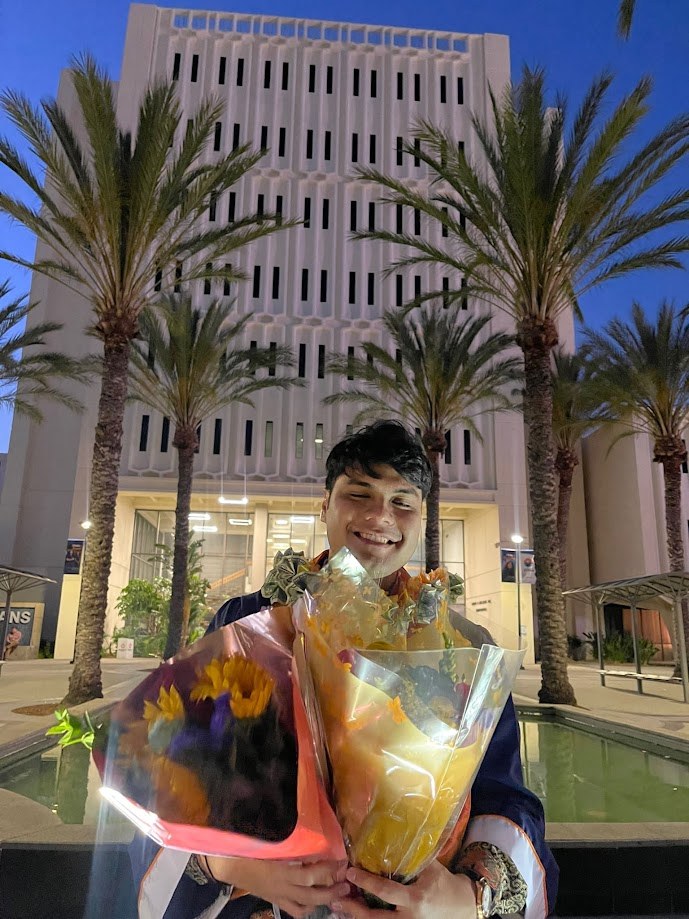 Holding flowers in cap and gown outside the CSUF library at dusk