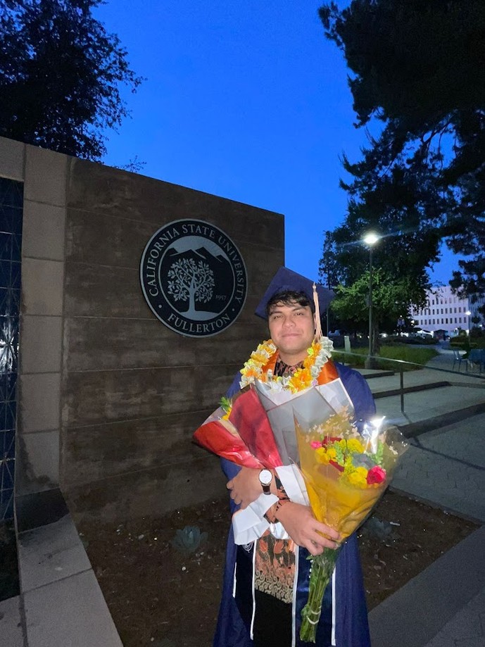 Holding flowers in cap and gown beside the Cal State Fullerton sign at dusk
