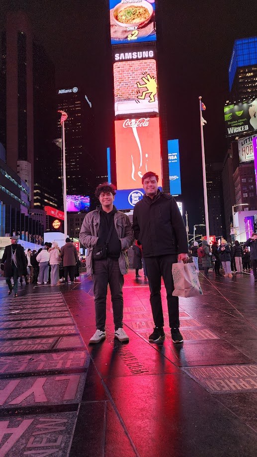 Two of us standing in the middle of Times Square at night, pavement wet