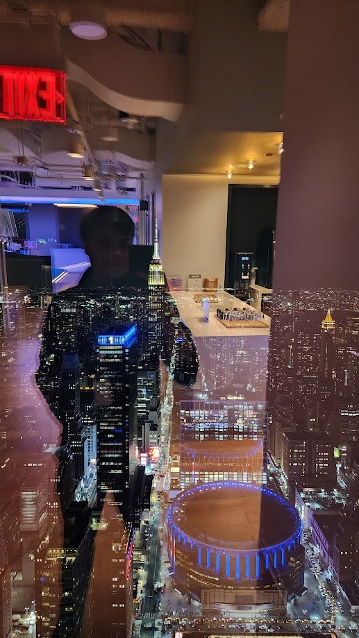 My reflection over the night view of Madison Square Garden lit blue