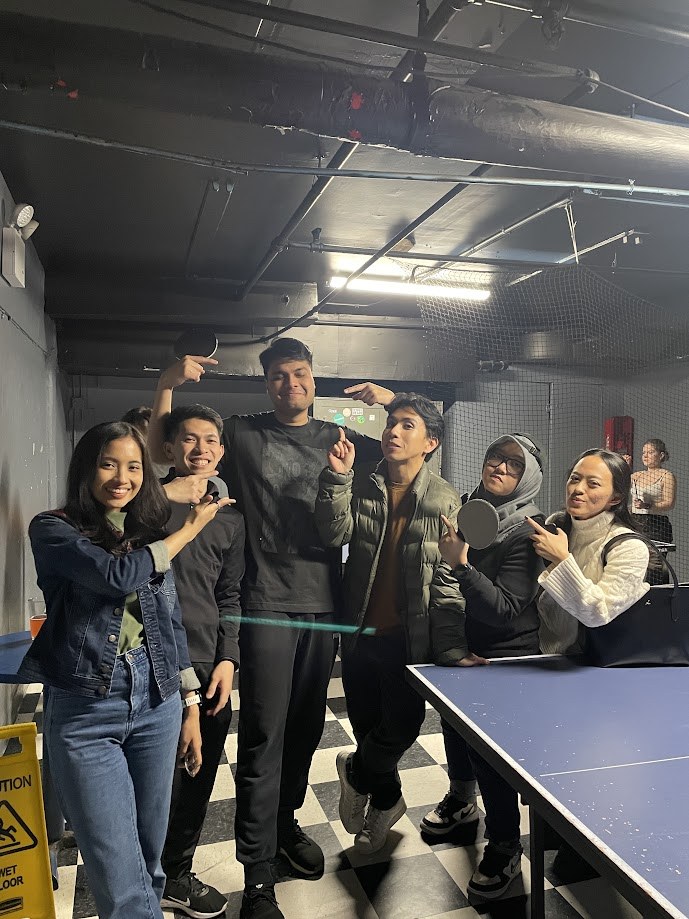 The group posing with paddles around a basement ping pong table