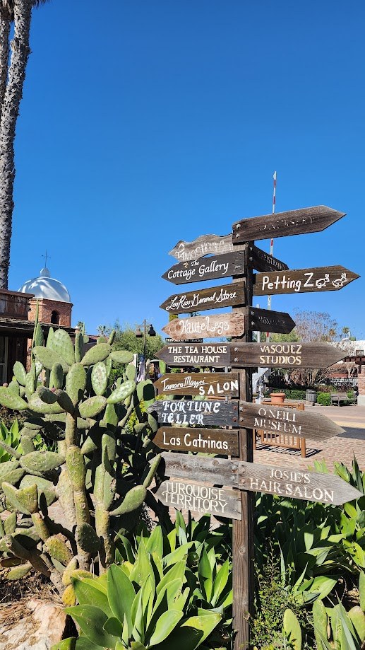 A wooden signpost pointing to the Petting Zoo, the Fortune Teller and the Cottage Gallery, cactus in the foreground