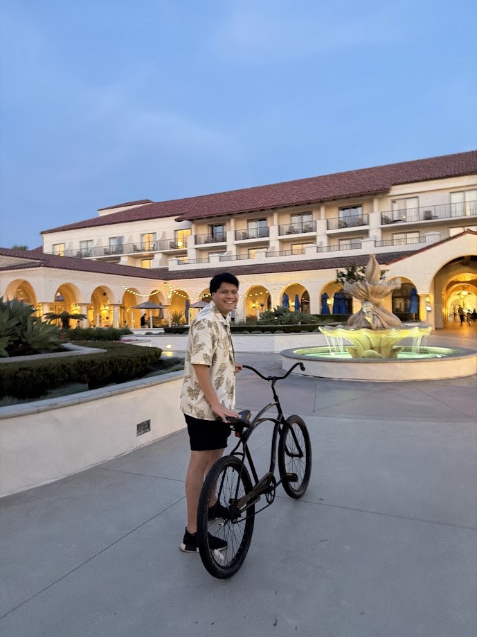 Standing with a beach cruiser in a lit courtyard by the fountain at dusk