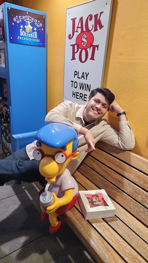 Leaning on a bench beside a Milhouse souvenir cup under a Jackpot sign